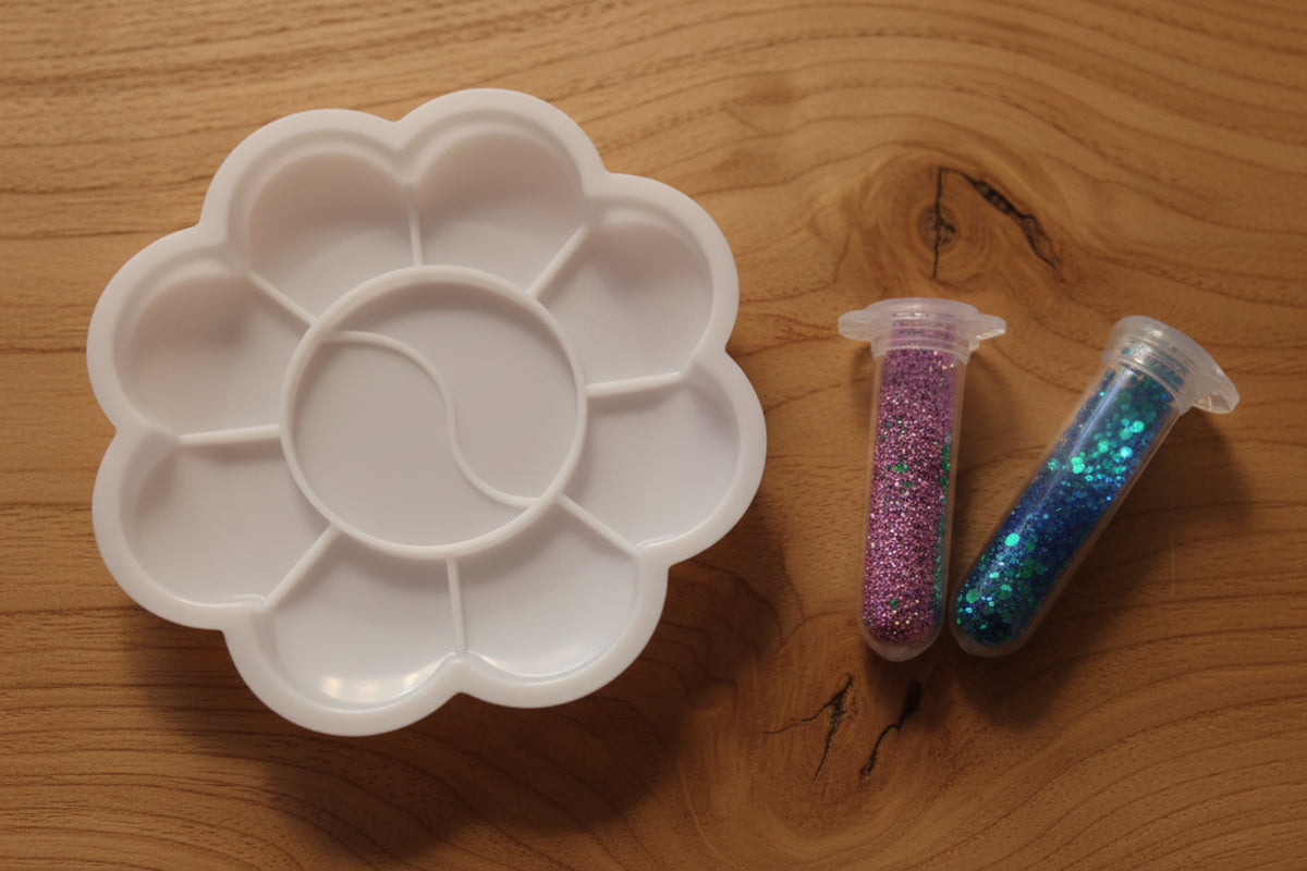 A white plastic flower-shaped paint palette with six compartments, shown on a beige fabric background with two small glitter tubes above it.